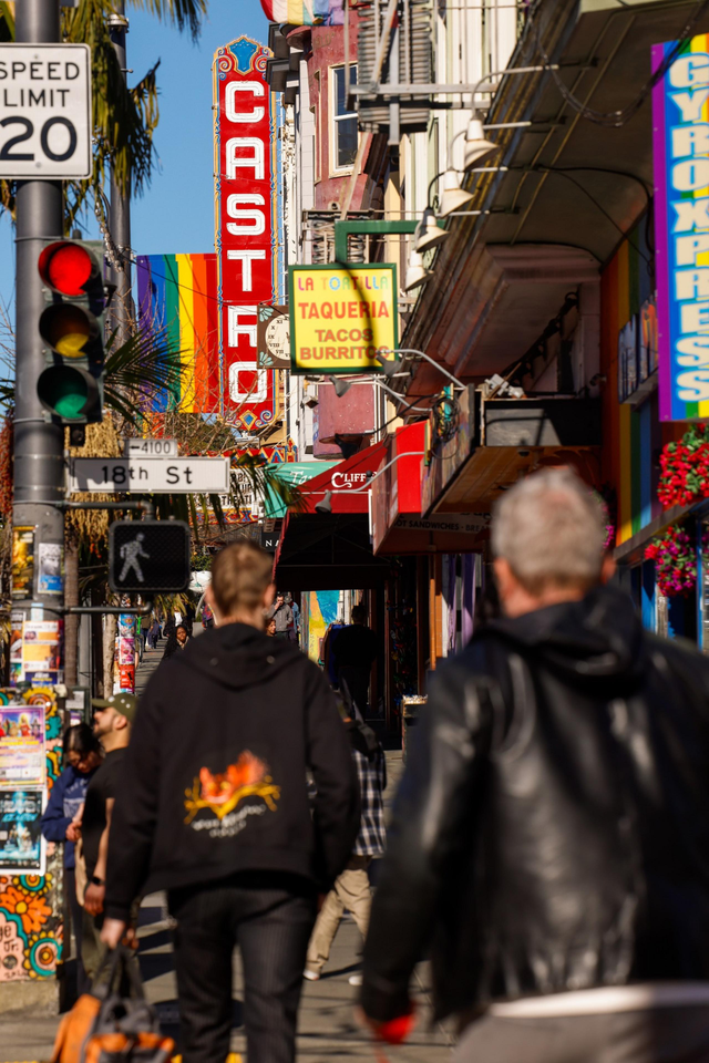 People walk along a vibrant street with colorful signs, including a large “Castro” sign and rainbow flag, surrounded by bustling shops and a traffic light.